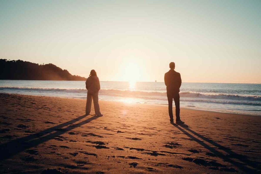 Two silhouettes standing apart on a beach at sunset, poem for him about one sided love and distance