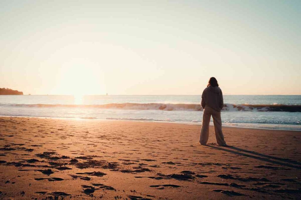 Woman standing alone on a beach at sunrise with footprints leading to the shore, poem for him about unrequited love