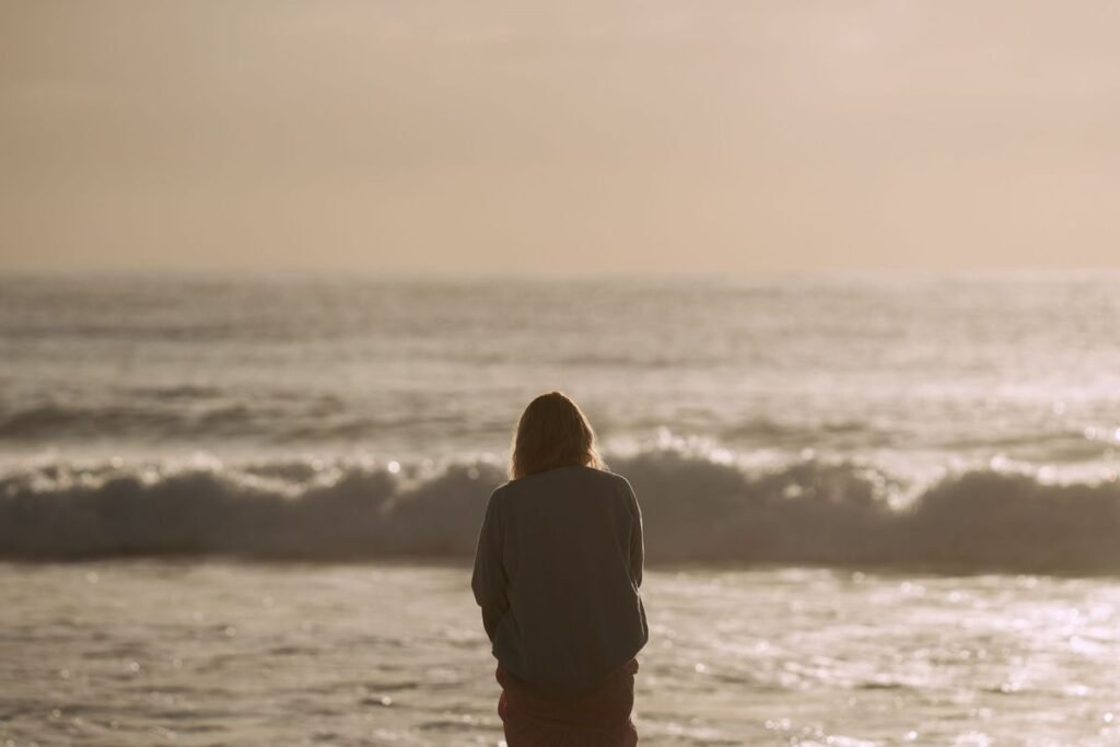 Woman standing alone facing ocean waves at golden hour, poem for him about lost love